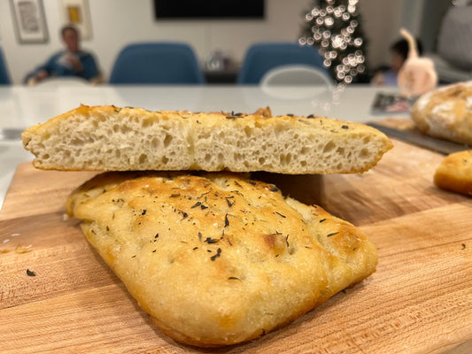 Crescent Bakes Focaccia bread topped with herbs, sliced, and stacked resting on a wood surface.