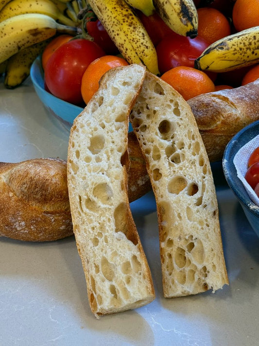 A Crescent Bakes Sourdough Baguette cut in half, displayed with a variety of fruits and other bread in the background.