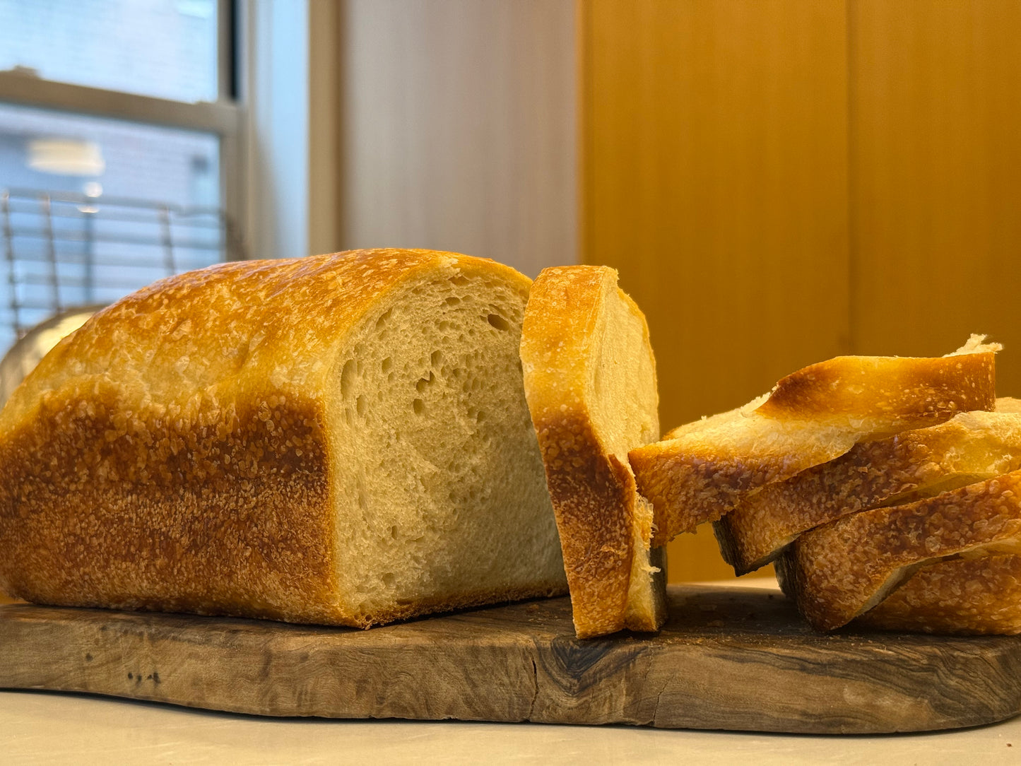 A partially sliced Crescent Bakes Sourdough Sandwich loaf on a wooden cutting board with a window in the background.