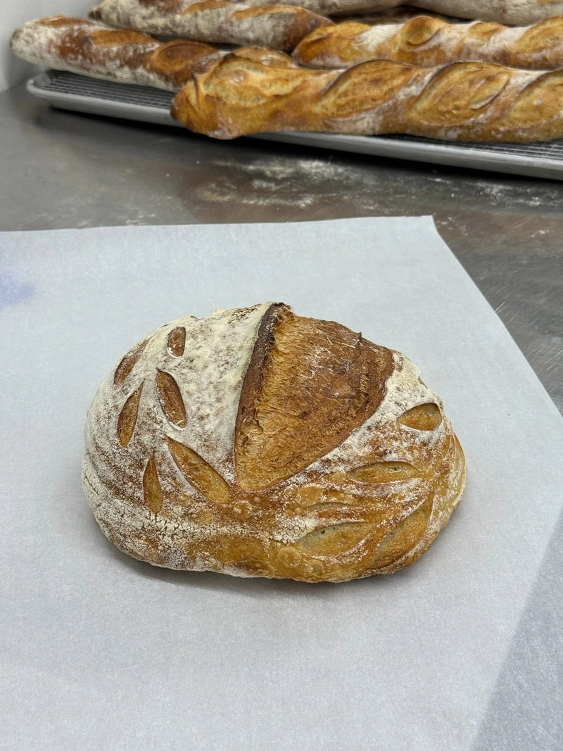 A freshly baked Crescent Bakes Sourdough Bread loaf on a wooden cutting board with a blurred kitchen background.