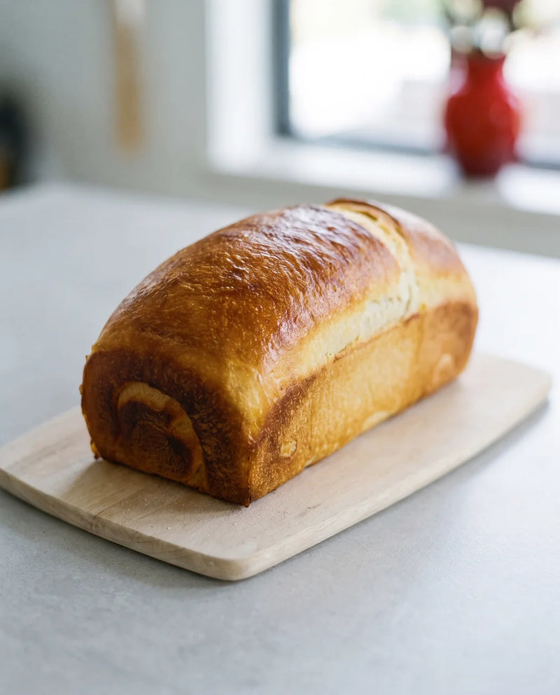 A partially sliced Crescent Bakes Sourdough Sandwich loaf on a wooden cutting board with a window in the background.